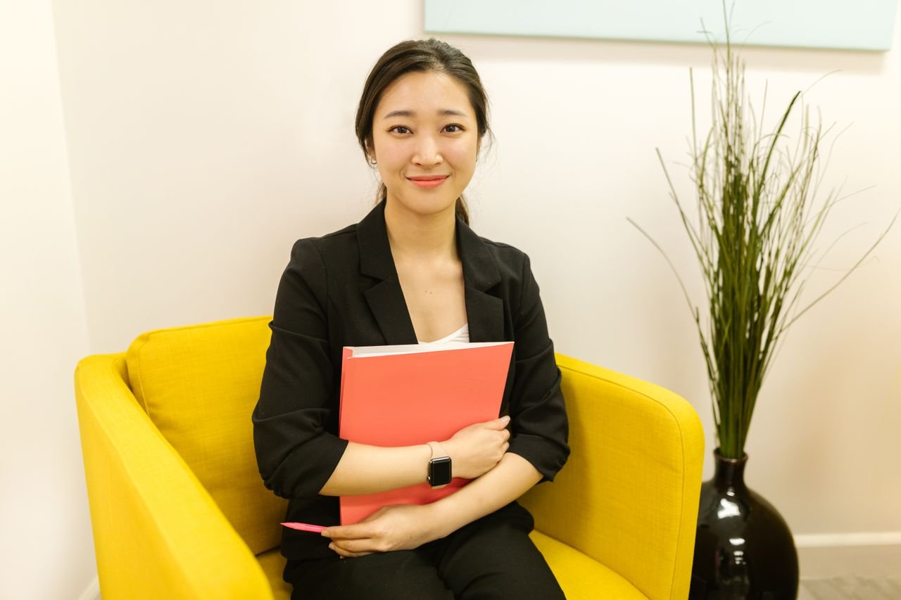 pexels photo 7845394 Elegant Asian woman smiling while holding a folder, seated in a modern office chair.